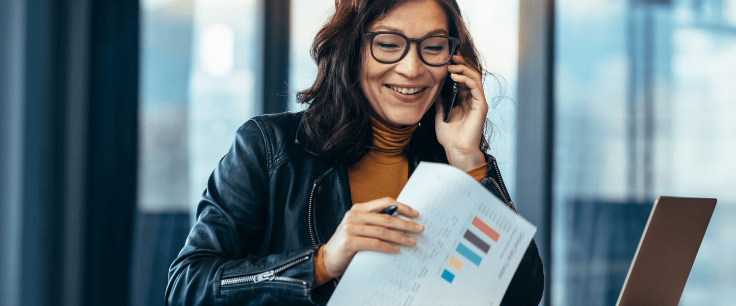 A woman with glasses smiling while talking on her phone and looking at a colorful report.