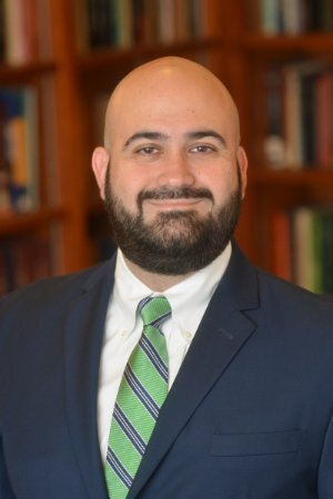 Dr. Oscar J. Mayorga standing in front of a bookshelf, wearing a dark suit and green striped tie.